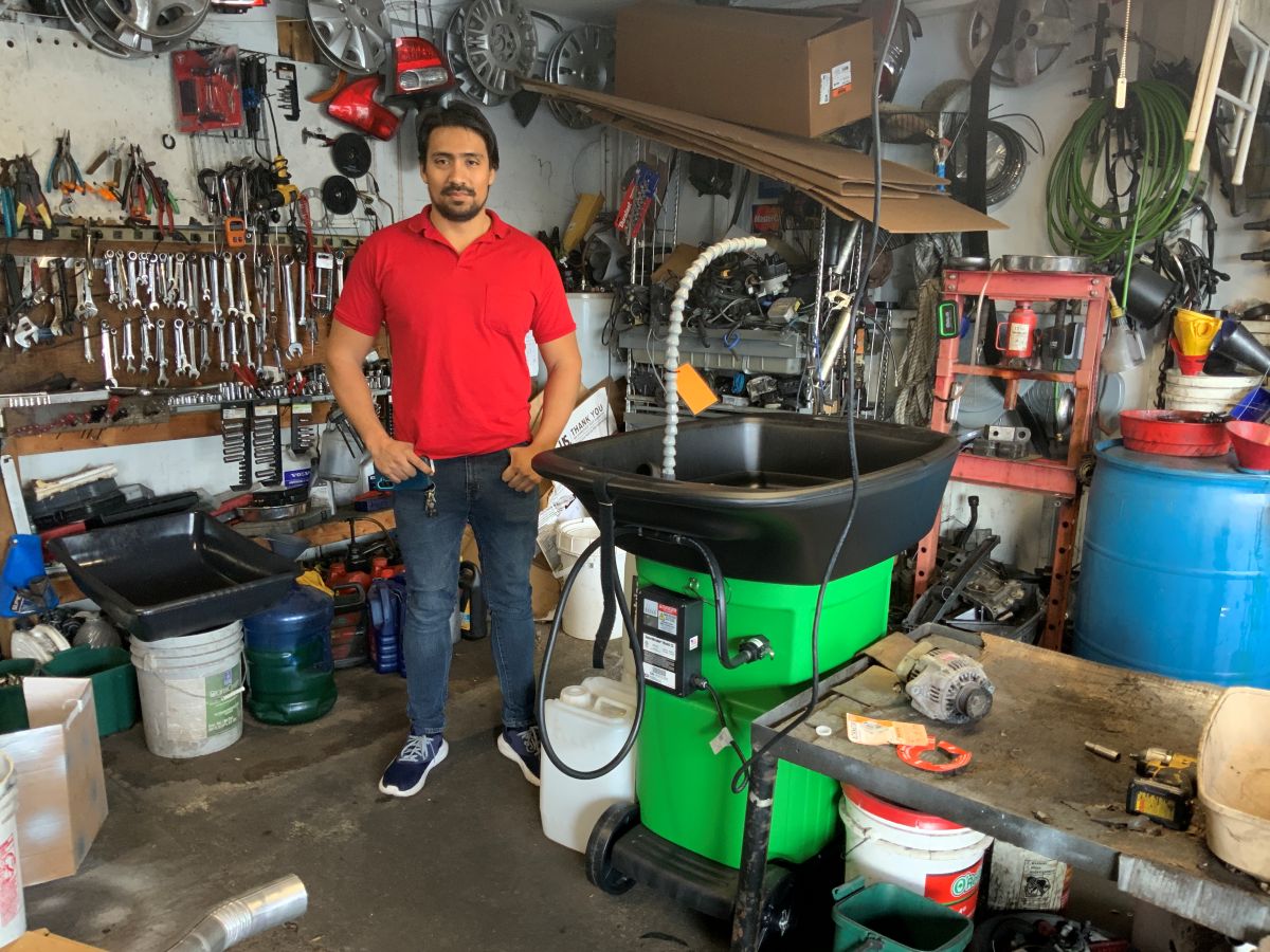 A man in a red shirt and blue jeans stands in an auto repair garage next to a bright green degreasing sink.