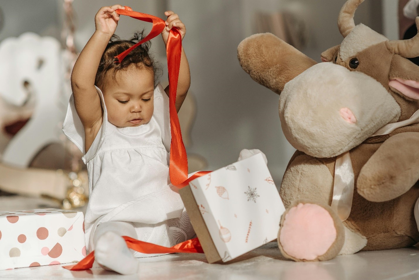 A child plays with a red ribbon coming out of a white wrapped present next to stuffed animals