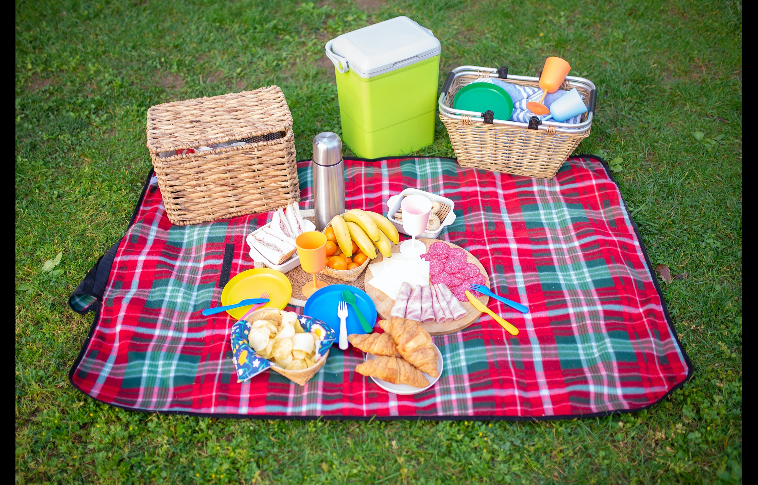 A red and green plaid blanket sits on a lawn and contains crackers, meats, cheese, and picnic baskets and coolers