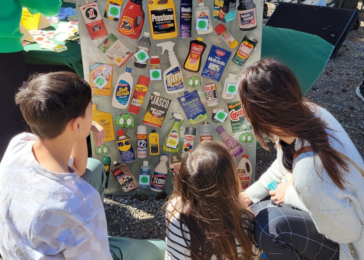 Three people at an outdoor event look at an educational display board filled with magnets resembling various cleaning product bottles.