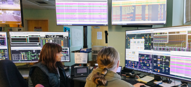 Two employees seated in a control room with numerous computer screens.