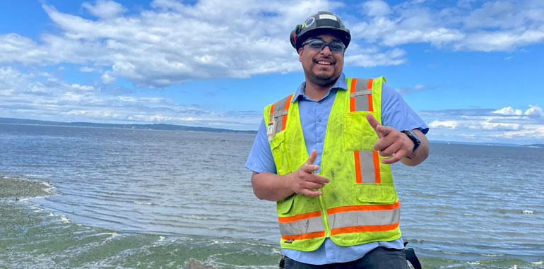 King County employee in bright yellow vest, hard hat and safety glasses standing on the beach next to the West Point Treatment Plant in Seattle.