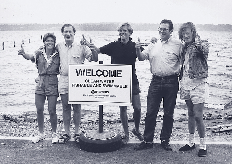5 people standing at the edge of Lake Washington next to a sign "Welcome. Clean Water, Fishable and Swimmable. Metro."
