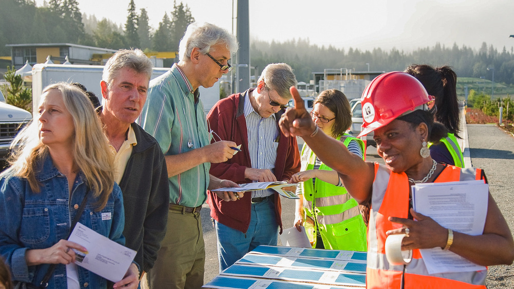 At Brightwater Treatment Plant, a group of adults at an outdoor information session, with a woman in a safety vest and hard hat gesturing and explaining, while attendees listen and hold papers.