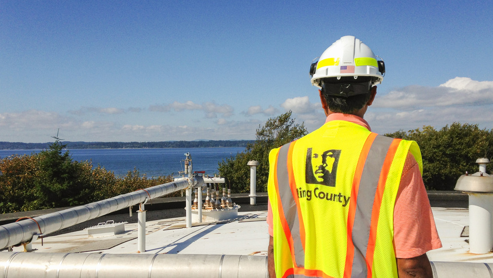 A person with safety equipment (hard hat and bright yellow vest) looks at the Puget Sound in the distance and the top of wastewater treatment tank in the foreground.