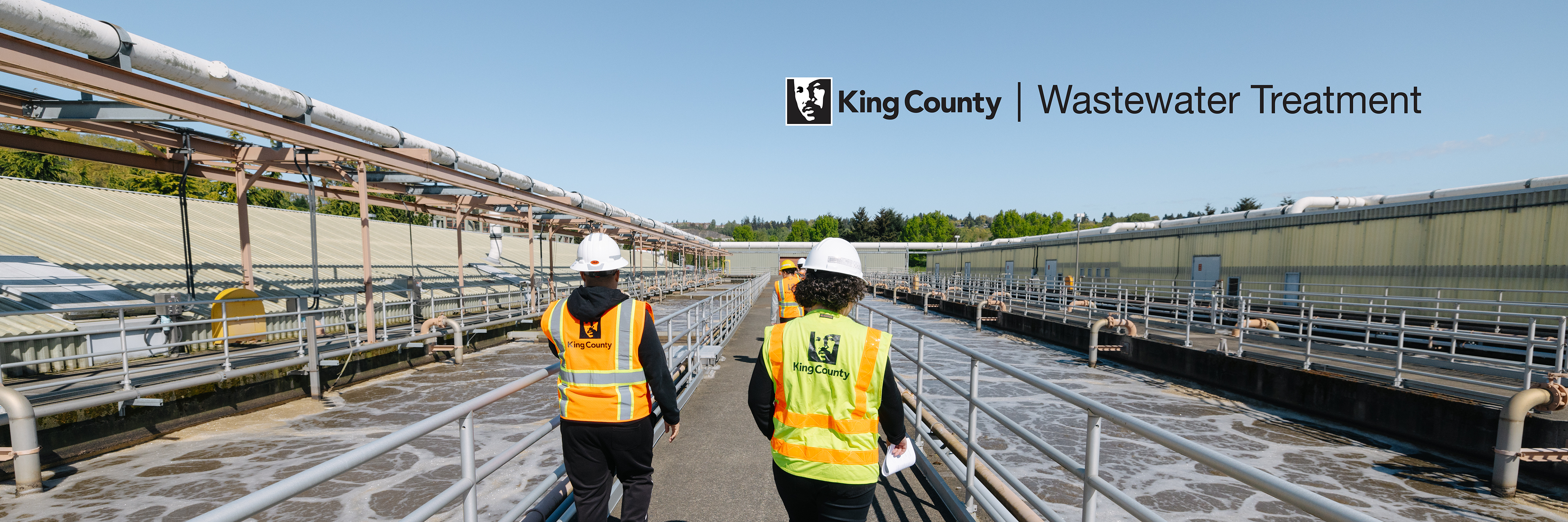 Three King County workers in safety gear (hard hat and yellow vests) walk along a pathway next to water being treated in tanks.