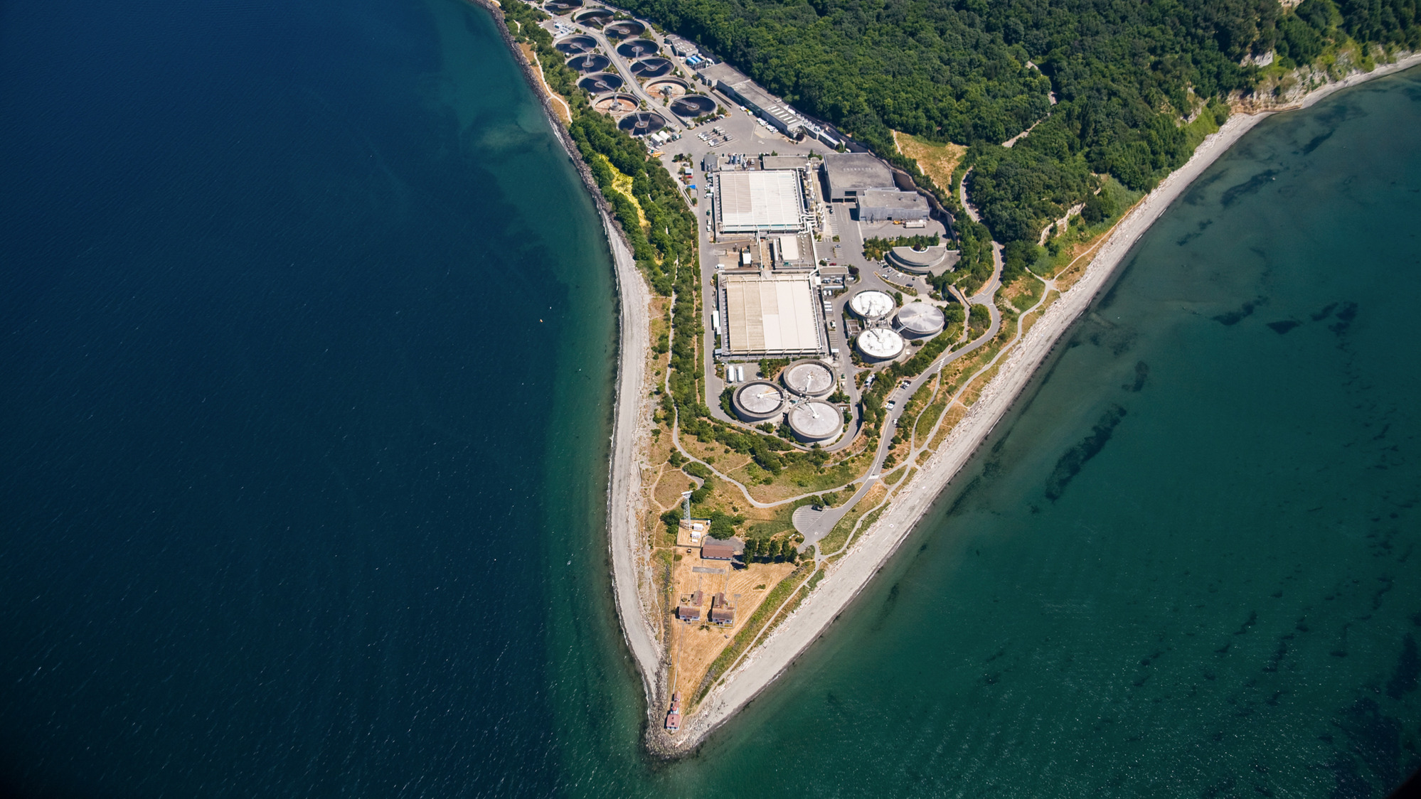 Aerial view of a West Point Treatment Plant located on a narrow strip of land between a body of water and a forested Discovery Park.