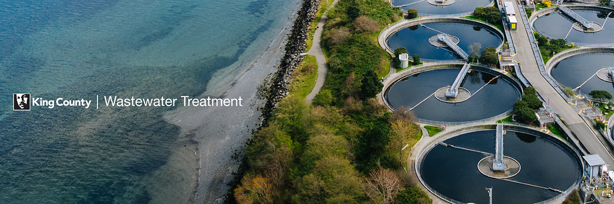 Aerial view of five circular tanks full of water going through the treatment process. The tanks are located close to a salt-water beach separated by a fenced area allowing public access along the beach. 