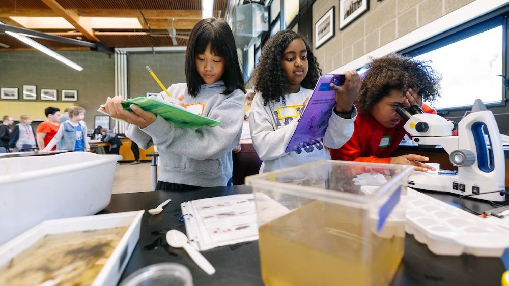 Two students are taking notes while a third student is looking through a microscope in a classroom at the Brightwater Education Center. 