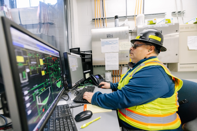 A wastewater operator, dressed in a yellow high-visibility vest and black hard hat, works on a computer. It shows graphs and charts about the operation of a wastewater facility.