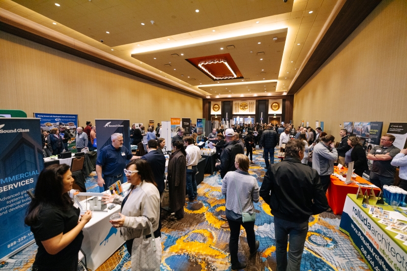 Busy networking event in a large conference hall, with rows of vendor booths, attendees talking, and bright overhead lighting.