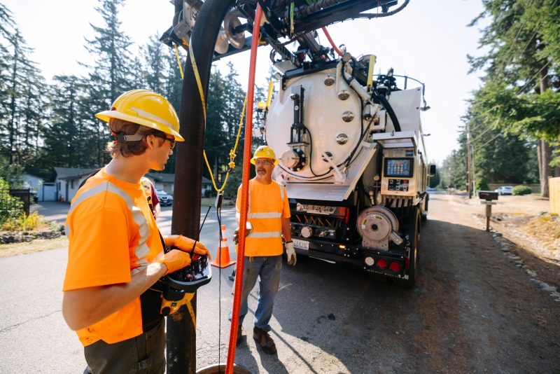 Two workers wearing orange high-visibility shirts and yellow hard hats operate a sewer cleaning truck on a street surrounded by trees.