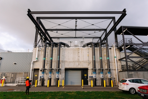 Exterior of the Georgetown Wet Weather Treatment Station. An industrial building with vertical piping from the ground up to the roof, a rollup entry door, and external stairway. An employee with hard hat and safety vest is standing outside. 