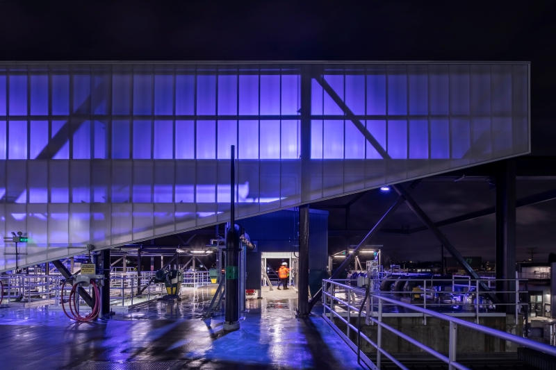 The image shows the outside of the Georgetown Wet Weather Treatment Station at night. The sky is fully dark, and the building is lit up with violet lights. 