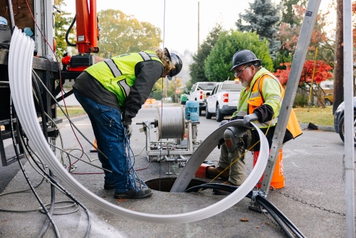 Two employees in personal protective gear are guiding a flexible tube into a maintenance hole. 