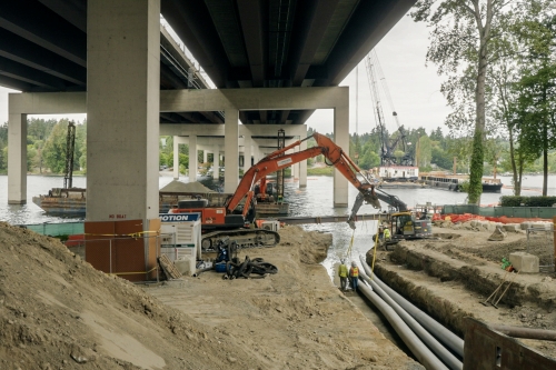 Under a highway overpass next to the water's edge, machinery is moving piping in a trench. 