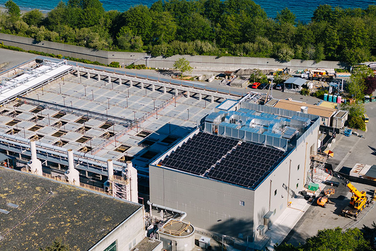 Aerial view of the Power Quality Facility at West Point Treatment Plant. The image shows a large building with solar panels on top, with trees and water in the background.