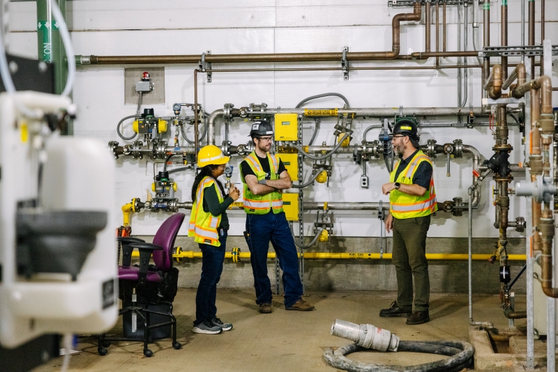 Three workers dressed in yellow high-visibility vests and yellow hard hats look at metal equipment inside of an industrial building. The equipment helps with the operation of a cogeneration facility.
