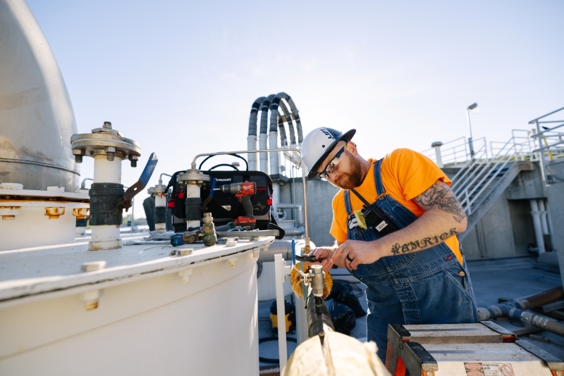 An operator dresses in an orange shirt, denim overalls, and a white hard hat fixes a piece of equipment on a sunny day with buildings at a treatment plant in the background.