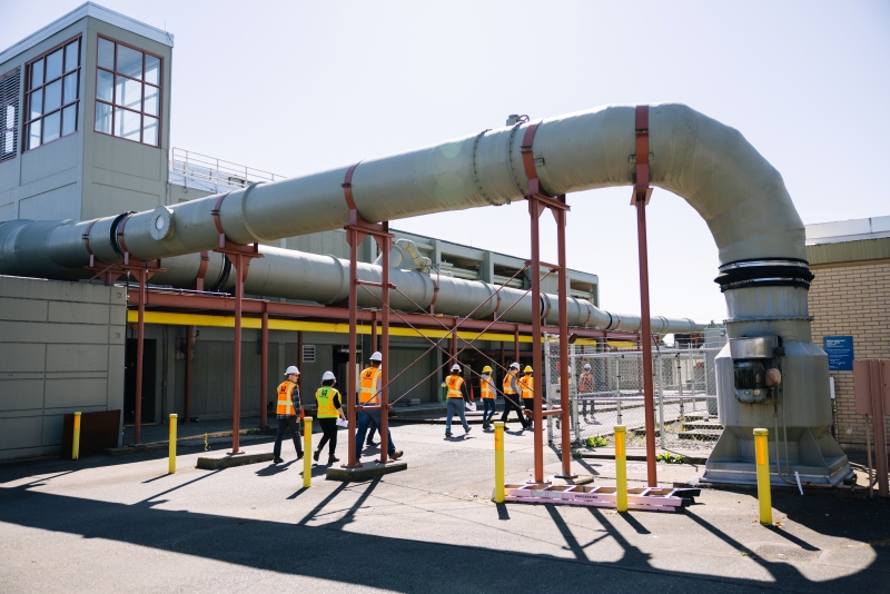 A group of people wearing orange high-visibility vests walks on a path outside at a treatment plant.