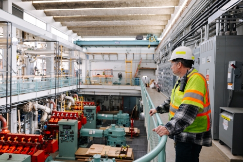 An employee in personal protective gear looks over a railing in an industrial room containing large pumps and mechanical equipment. 