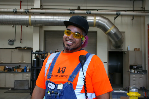 King County employee wearing an orange work shirt with reflective tape, safety glasses and ear protection in an industrial workshop. 