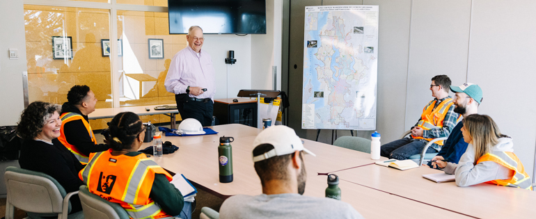 A group of people at a classroom table with an instructor standing at the head of the table. A large poster displays the King County wastewater system. 