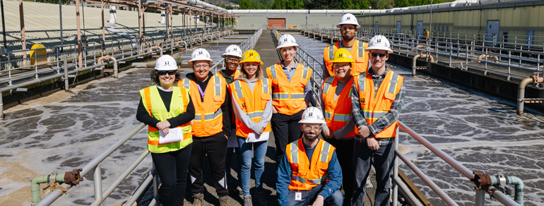 Workforce Development "cohort" - a group of nine people wearing safety gear (orange reflective vests, hard hats, eye protection). In the background are two tanks holding water flowing through the treatment process. 