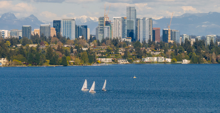 A photo showing three sailboats in a body of blue water in the foreground. The background is a tree lined shoreline, a city skyline, some snow capped mountains and a cloudy sky.
