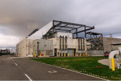 A large industrial facility with pipes, metal framework, and utility infrastructure stands beside a paved roadway under an overcast sky.