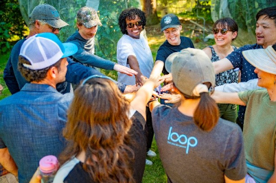A group of people standing in a circle extend their hands toward the center in an outdoor setting surrounded by trees.