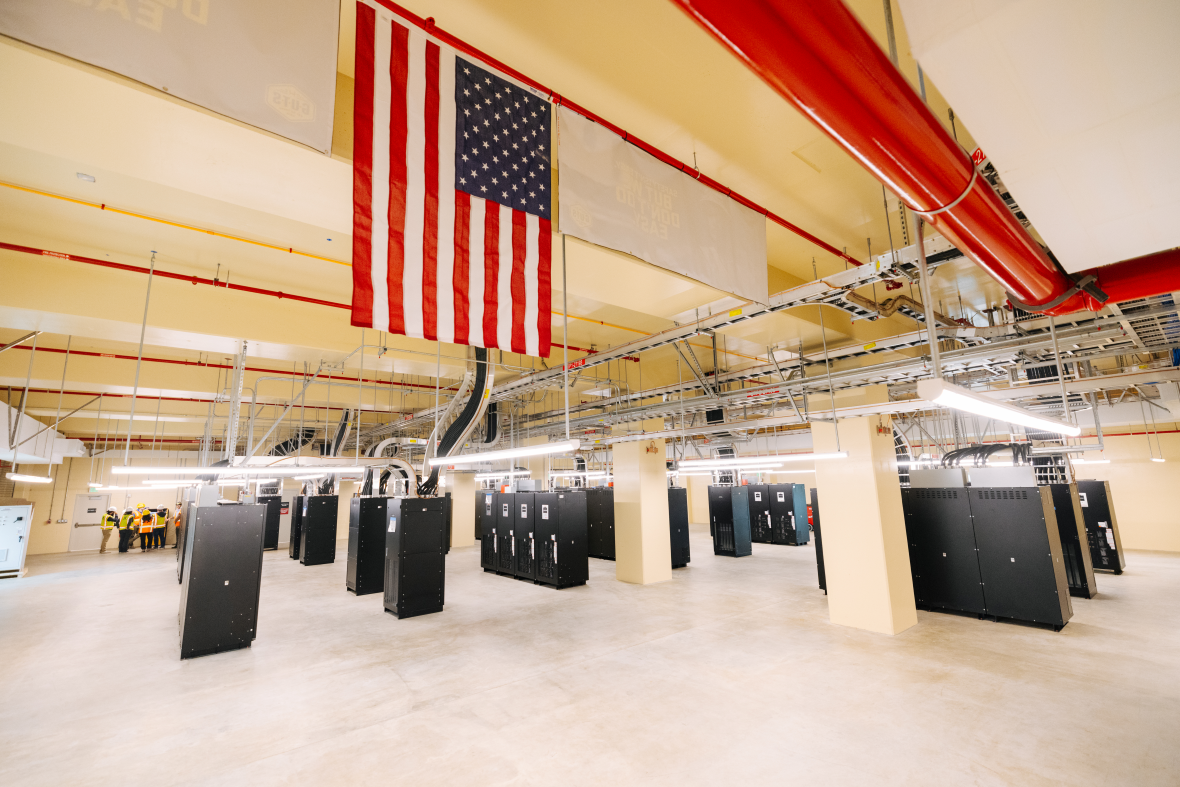 A large American flag hangs above rows of tall black equipment cabinets in a brightly lit industrial room with exposed pipes and wiring overhead.