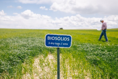 A blue sign reading “BIOSOLIDS 3 dry tons / acre” stands in the foreground of a green field with an individual walking in the background under a partly cloudy sky.