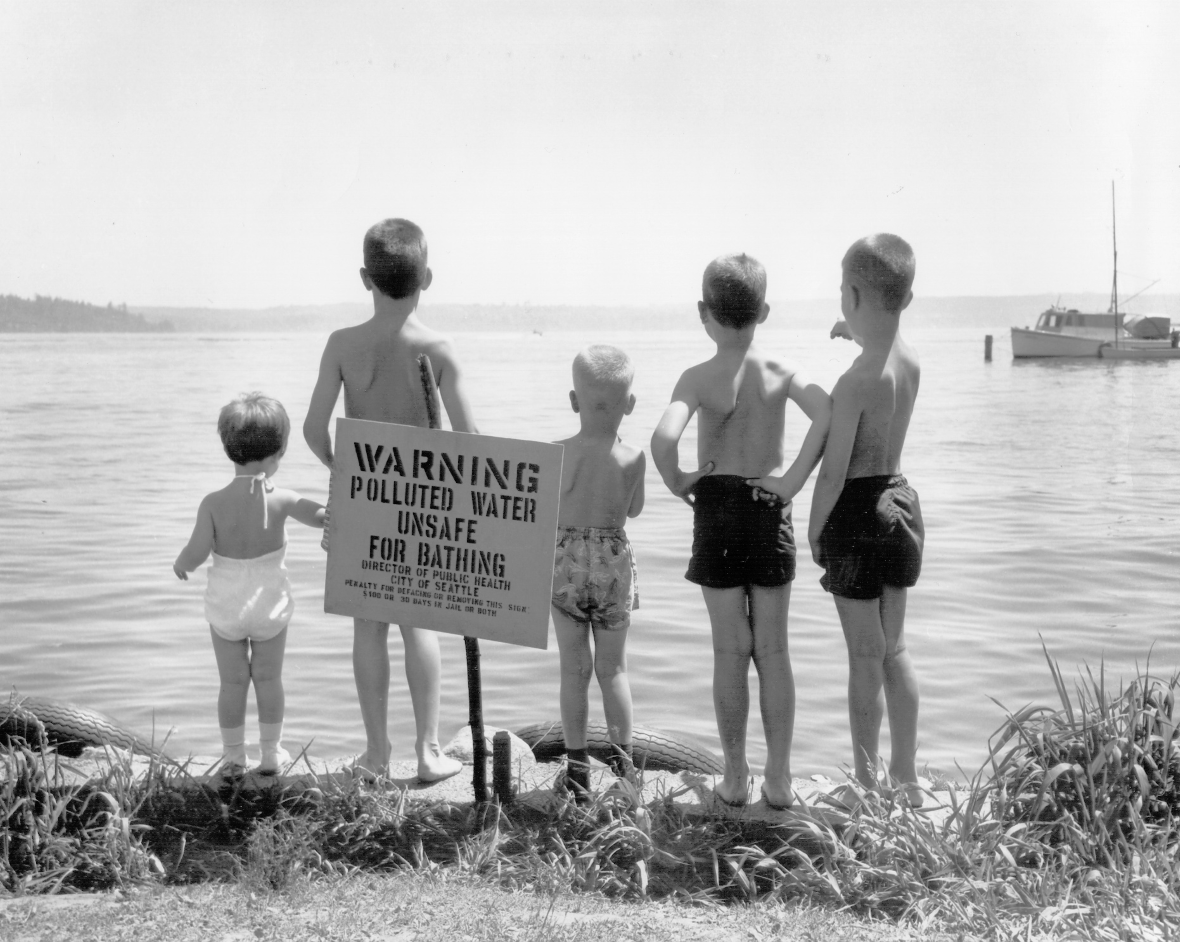 Five individuals stand at the edge of a shoreline beside a sign warning of polluted water, looking out toward the water and a boat in the distance