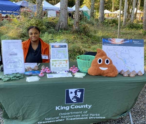 A King County representative is sitting behind a table with a green table covering with the King County Logo and "Department of Natural Resources and Parks, Wastewater Treatment Division" underneath. On the table are brochures and communication materials about the Black Diamond Sewer Upgrade project. 