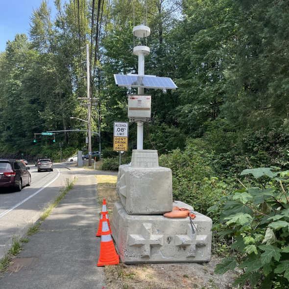 An electrical device with a solar panel on top of a concrete block, next to a sidewalk and street