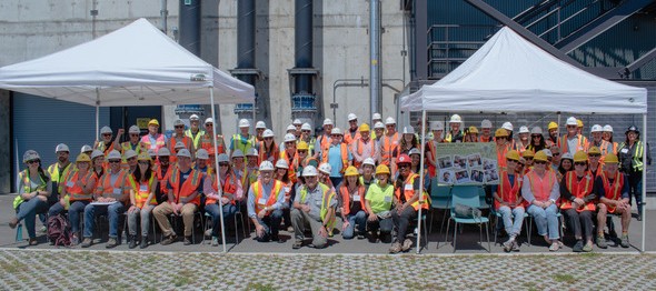 Project team in orange vests, glasses, and hard hats, posing under two white canopies in front of the Georgetown Wet Weather Treatment Facility