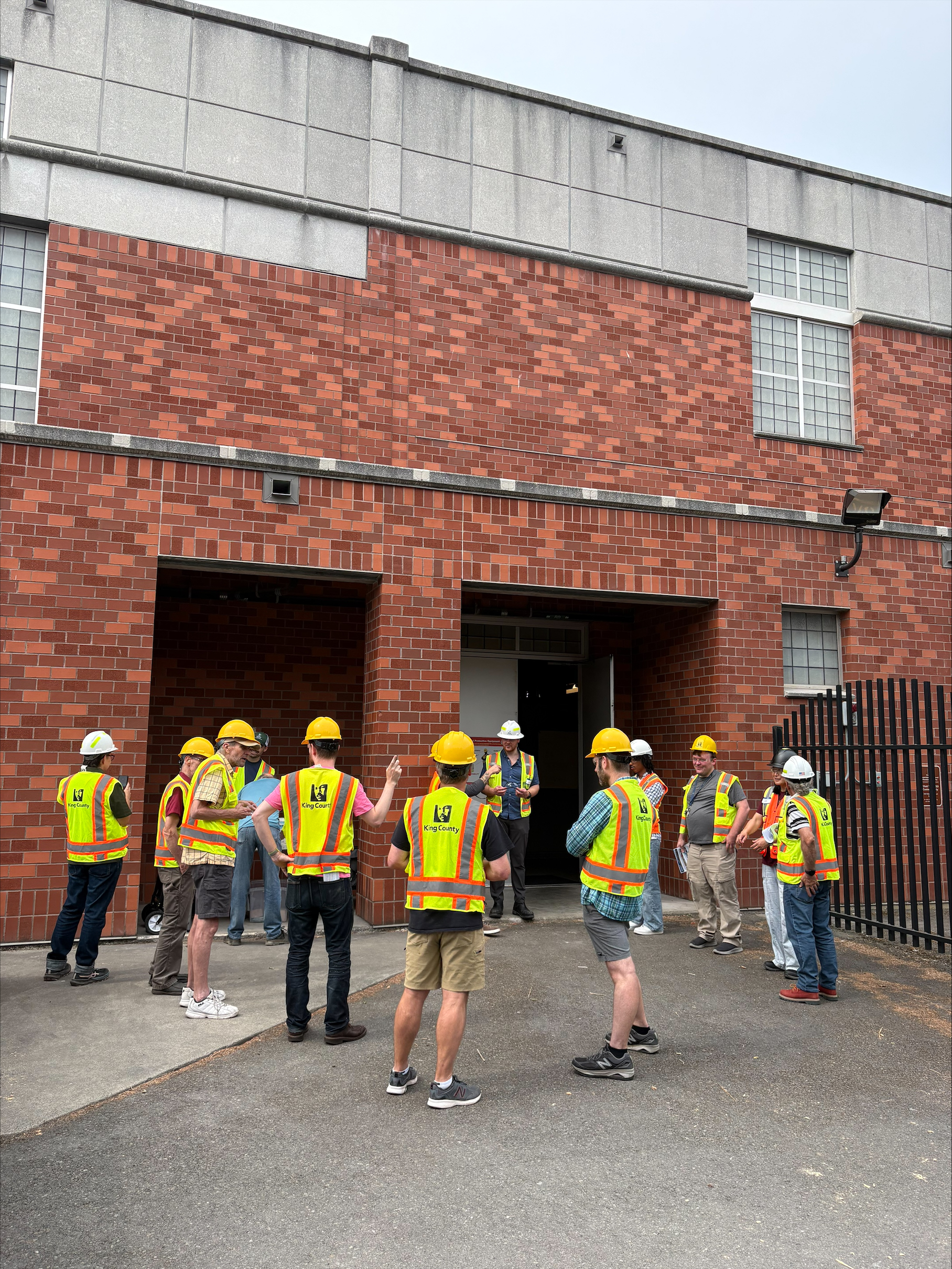  A group of community members and King County staff in yellow hard hats and safety vests gather in a semicircle outside the brick EWWTS facility, listening to a guide during the August 21 open house tour.