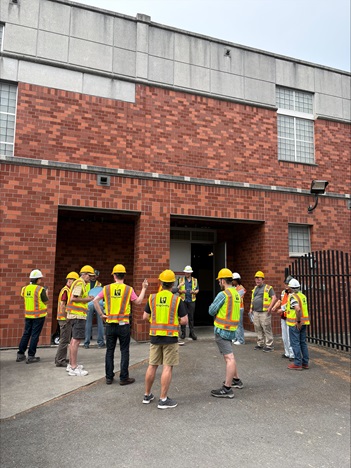 A group of community members and King County staff in yellow hard hats and safety vests gather in a semicircle outside the brick EWWTS facility, listening to a guide during the August 21 open house tour.