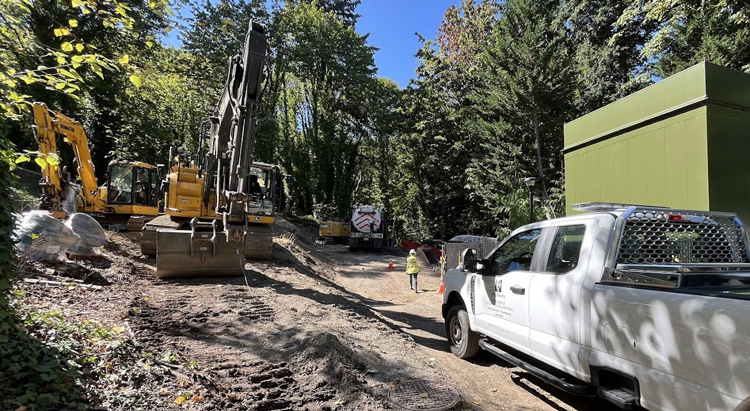 A worker walking through a construction area with excavators, a truck, and other construction equipment around. 