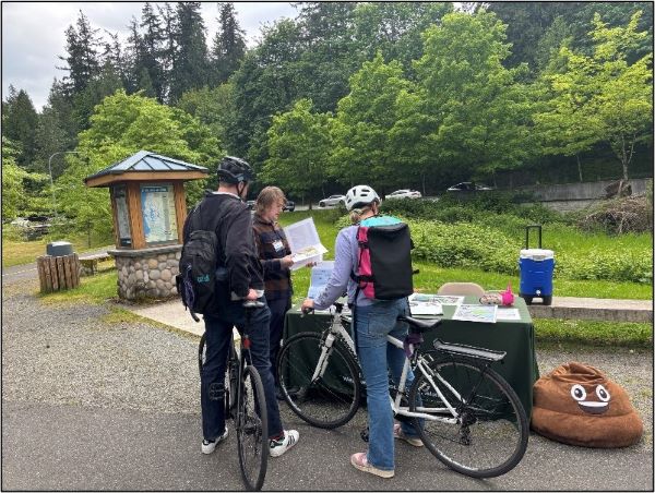 Two people with bicycles are next to a table on a pathway. A King County staff person is sharing some information about the sewer improvement project. 