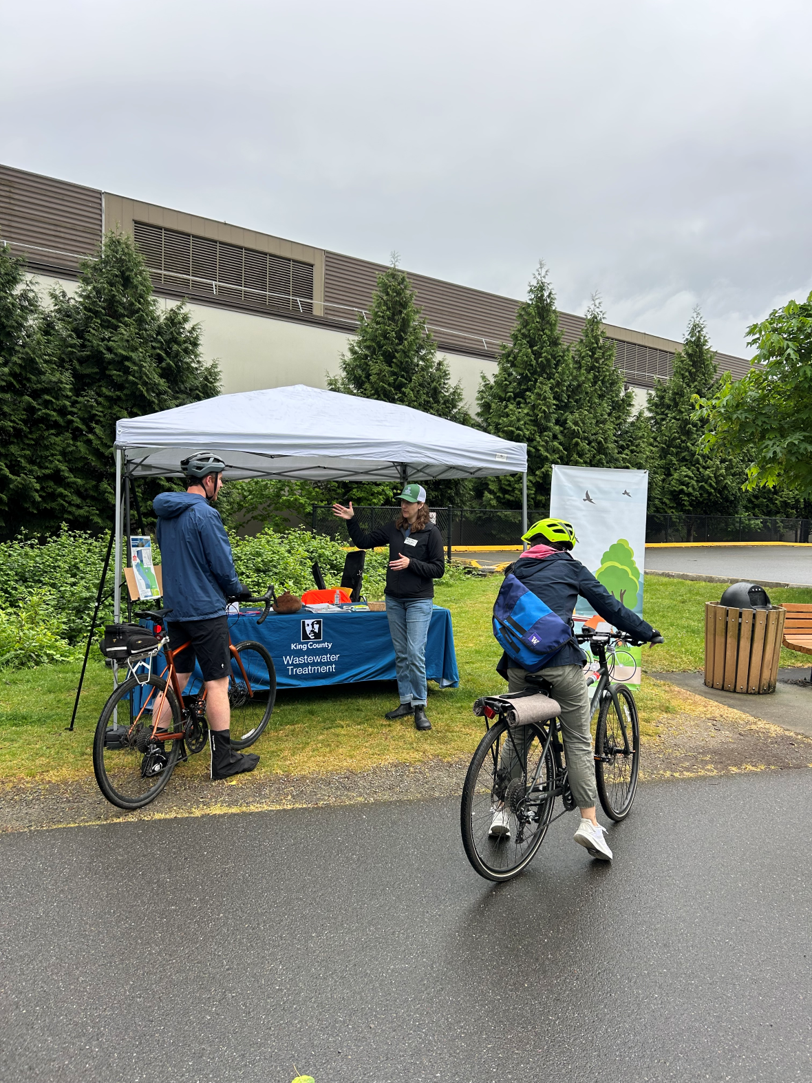 Cyclists stop at a King County Wastewater Treatment information booth under a canopy on a rainy day along a paved trail