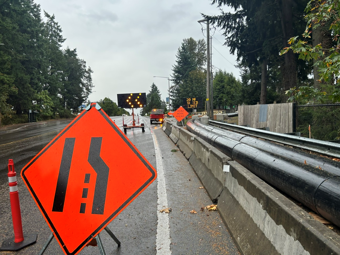 The image shows a rainy roadway with lane‑shift warning signs, cones, and an arrow board directing traffic through a construction area.