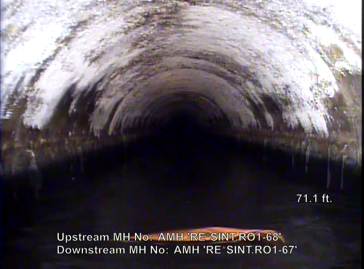 he image shows the inside of a large underground pipe with water at the bottom and deposits along the upper surface, captured from a sewer inspection camera.