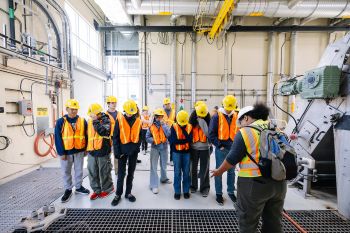 Group of middle school students wearing hardhats and safety vests looks at equipment at Brightwater Treatment Plant. Teacher is explaining trash removal process.