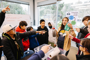 Teacher with group of youth. Teacher is explaining an experiment. There is an engineering design process poster on the wall.