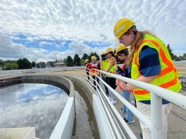 A group of the interns observe and take notes at one of the South Treatment Plants clarifiers. They stand along the rails of the clarifier, which is full of water and reflects the overcast sky that makes up the background. The students are wearing safety vests and hardhats.