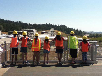 a tour group from elementary grades lined up along a railing with orange vests, hard hat and eye protection. 