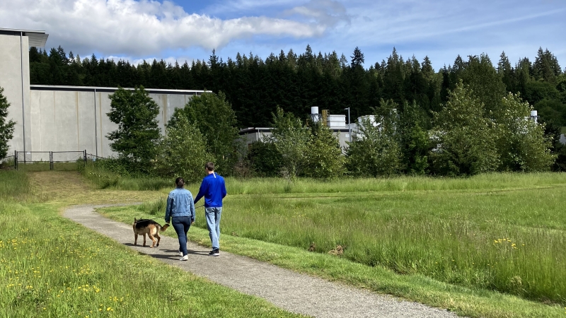 Two adults in blue shirts walk their leashed dog on a gravel trail. A concrete building is behind a fence in the background.