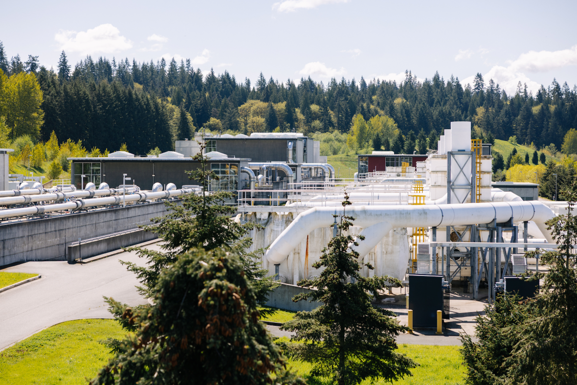 Looking out at an industrial facility with pipes in the foreground and large buildings in the background. Trees surround the facility.